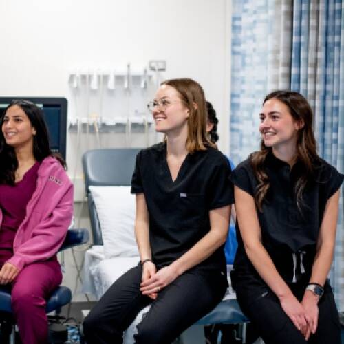 Three smiling students in scrubs sitting together in a clinical training room